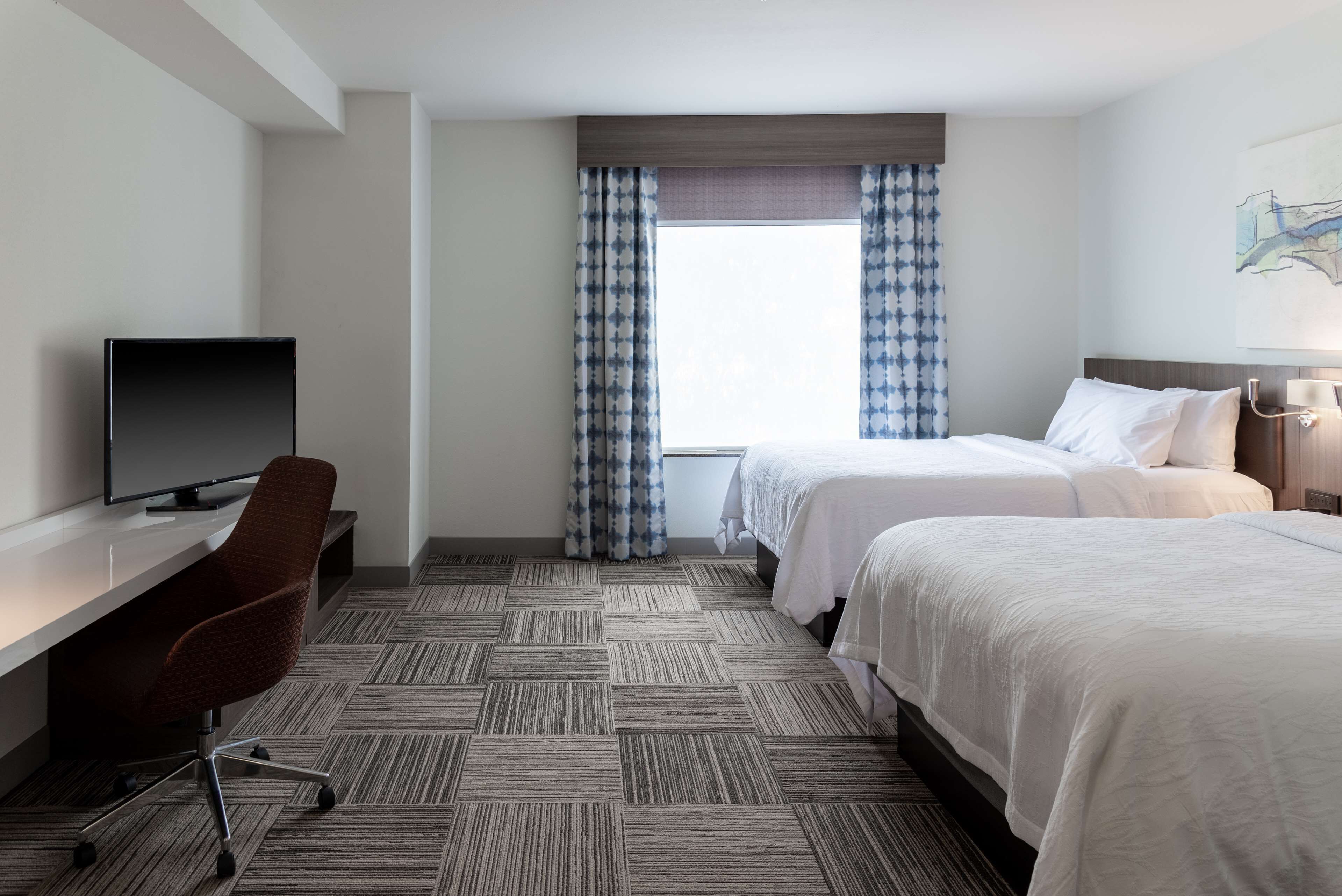 A hotel room with two beds, a desk with a chair, a television, and a window with curtains.