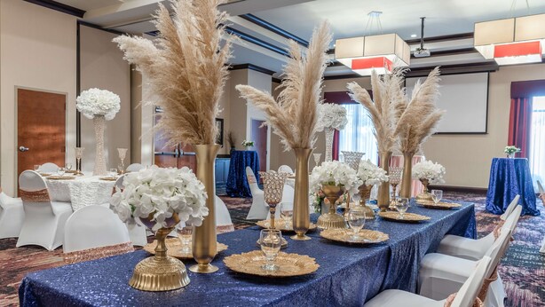 A banquet hall with tables set for a formal event, featuring white chairs, gold and white floral centerpieces, and a blue tablecloth. - Hilton Garden Inn North Little Rock (North Little Rock)
