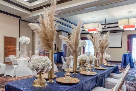 A banquet hall with tables set for a formal event, featuring white chairs, gold and white floral centerpieces, and a blue tablecloth.