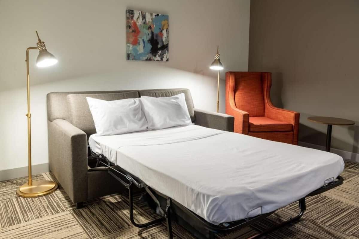A single bed with a grey headboard, two bedside lamps, a small orange chair, and a small round table.