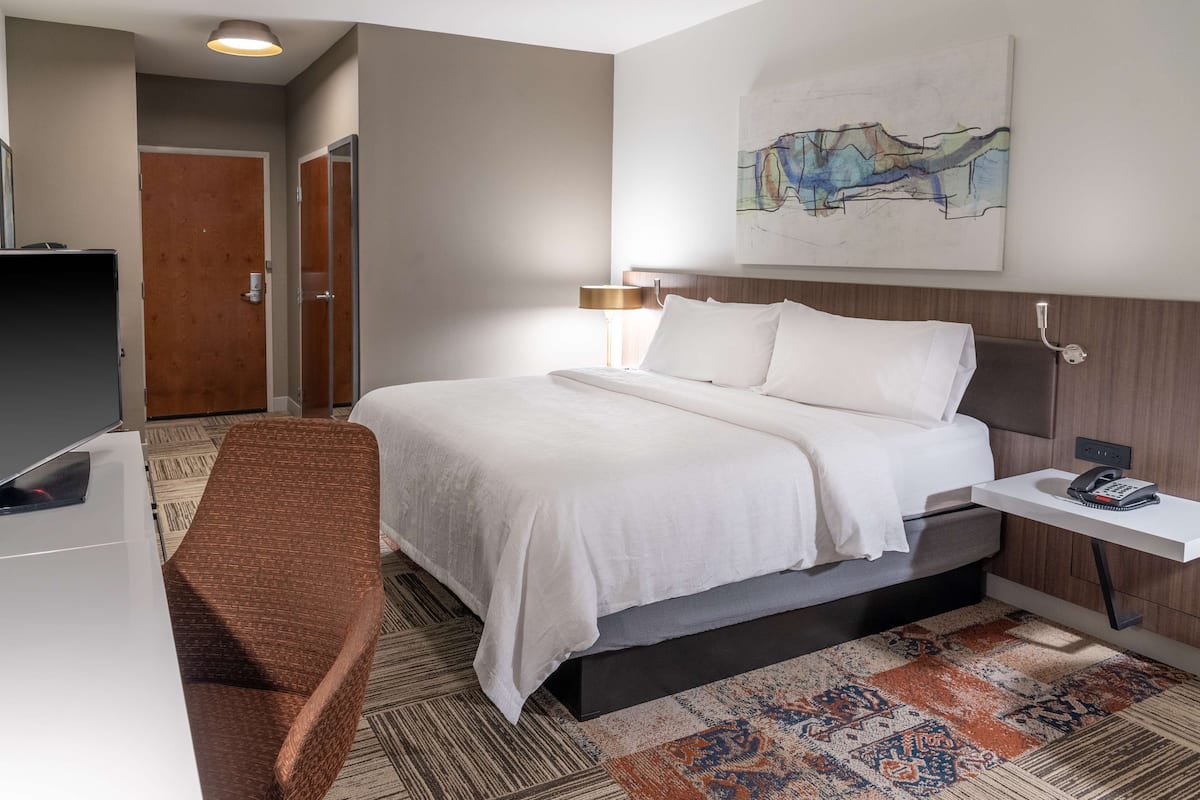 A hotel room with a bed, bedside table, lamp, and a painting on the wall.