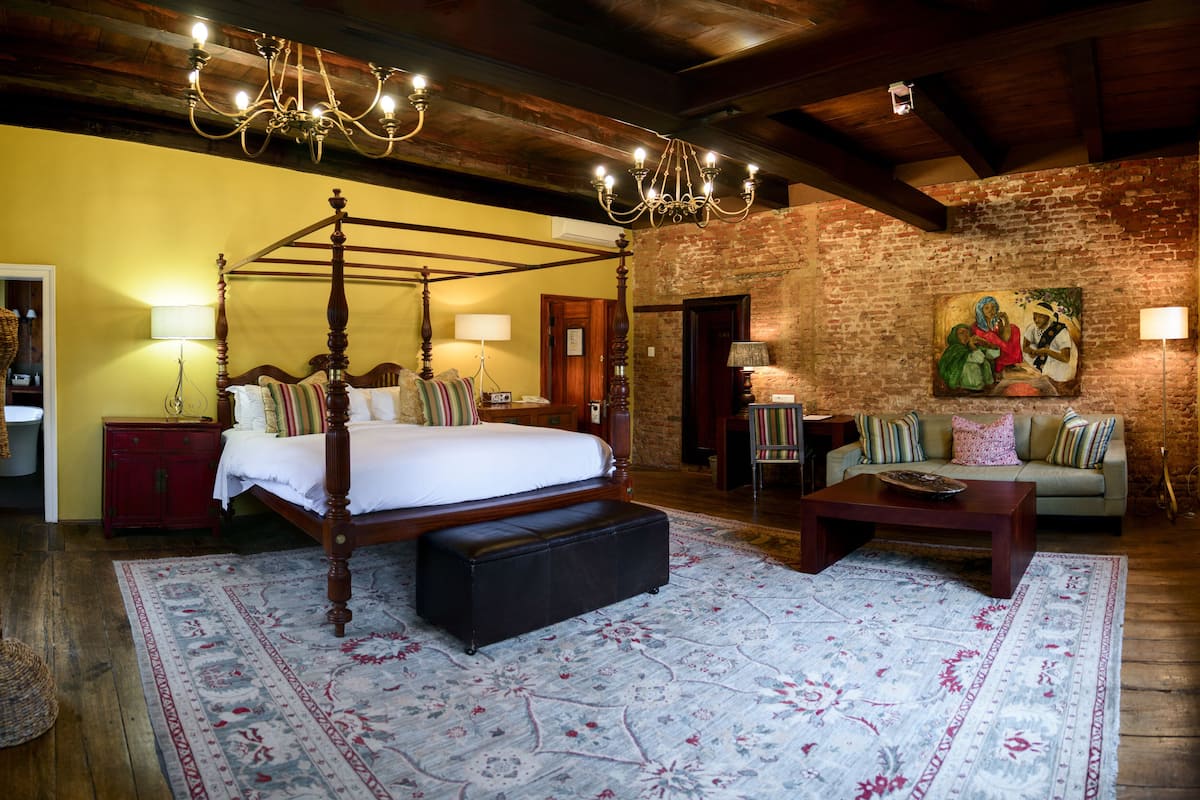 A four-poster bed with a bench, a wooden coffee table, and a patterned rug in a room with exposed brick walls and wooden beams.