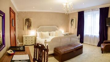A bedroom with a large bed, a wooden nightstand, a desk with papers, a leather ottoman, and a chandelier.