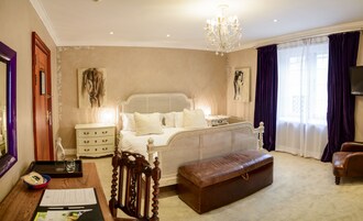 A bedroom with a large bed, a wooden nightstand, a desk with papers, a leather ottoman, and a chandelier.