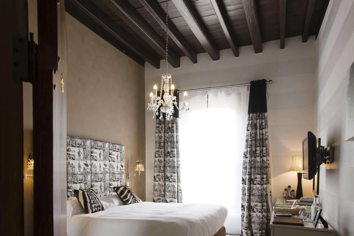 A bedroom with a large bed, a chandelier, a nightstand, and a view of the outdoors.