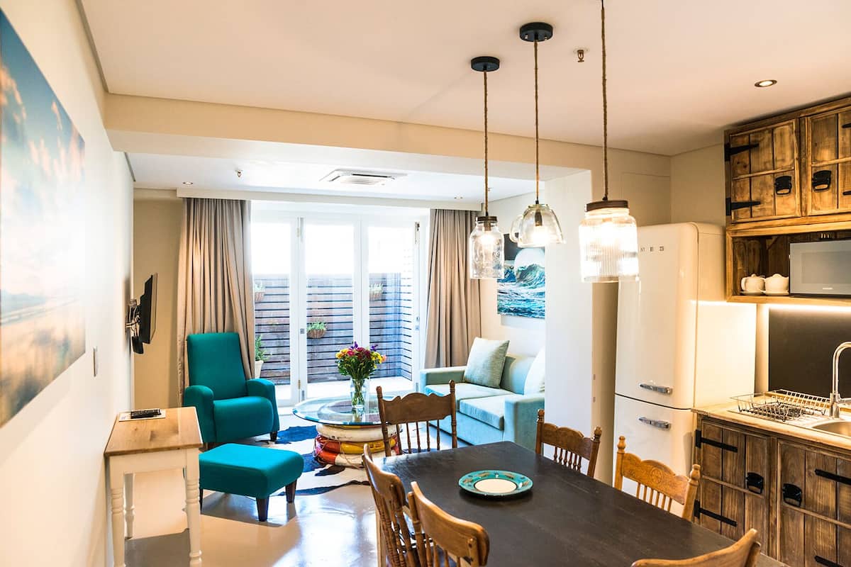 A modern kitchen with a refrigerator, microwave, and sink. There is a dining area with a table and chairs, and a living room with a sofa and a chair.