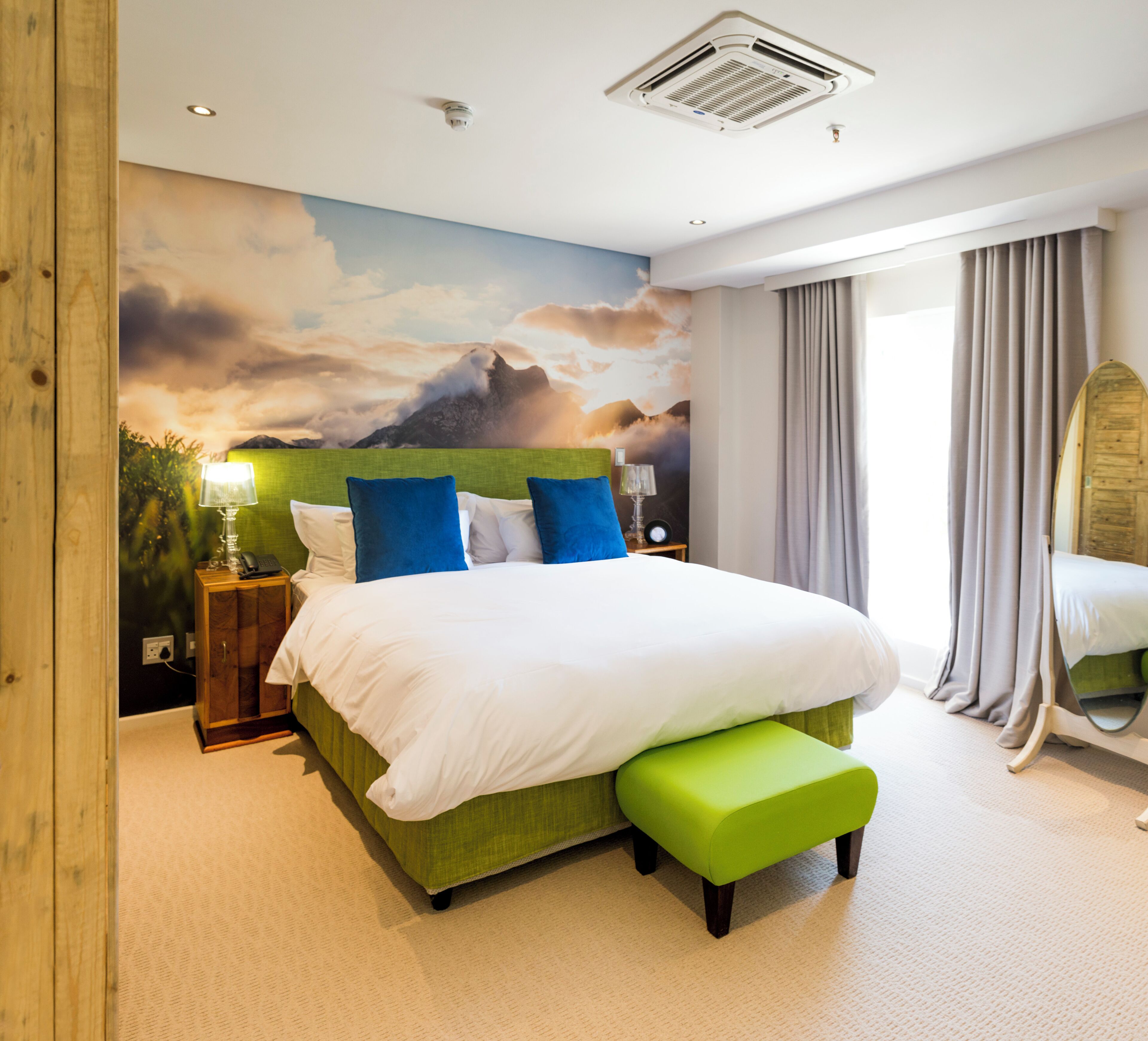 A bedroom with a bed, a green upholstered bench, a nightstand with a lamp, and a wall mural.
