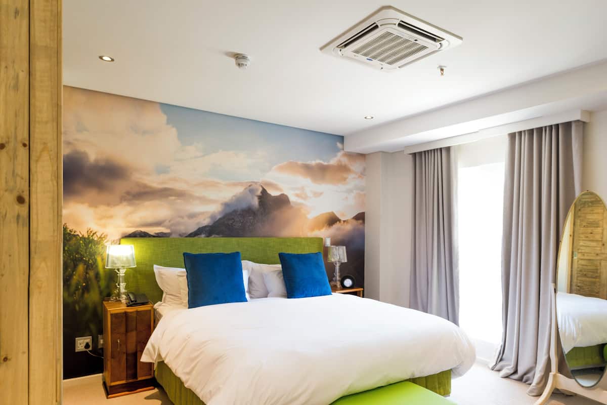 A bedroom with a bed, a green upholstered bench, a nightstand with a lamp, and a wall mural.