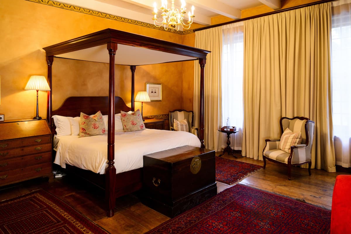 A four-poster bed with a wooden headboard, a red patterned rug, a wooden dresser, a chair, and a chandelier.