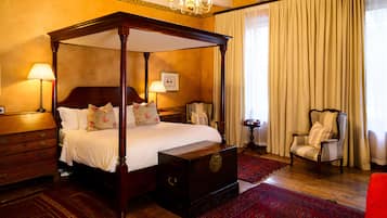 A four-poster bed with a wooden headboard, a red patterned rug, a wooden dresser, a chair, and a chandelier.