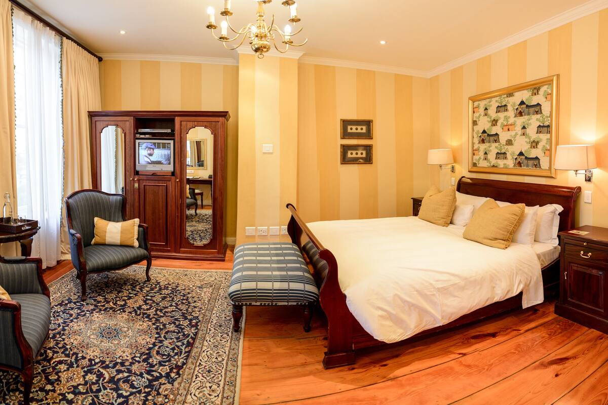 A hotel room with a large bed, a sitting area with a chair and ottoman, a wooden wardrobe, a television, and a painting on the wall.