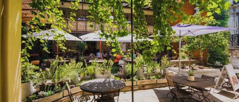 Un patio al aire libre con mesas y sillas, rodeado de plantas en macetas y sombrillas.