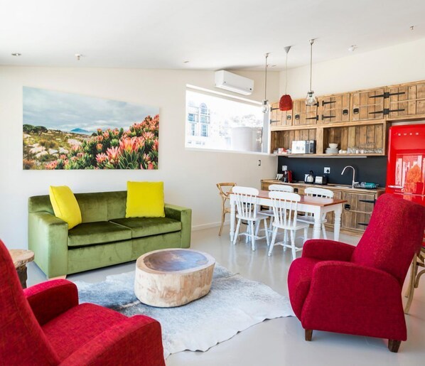 Una sala de estar moderna con un sofá verde, sillas rojas, una mesa de comedor y una zona de cocina con armarios de madera y un refrigerador rojo.