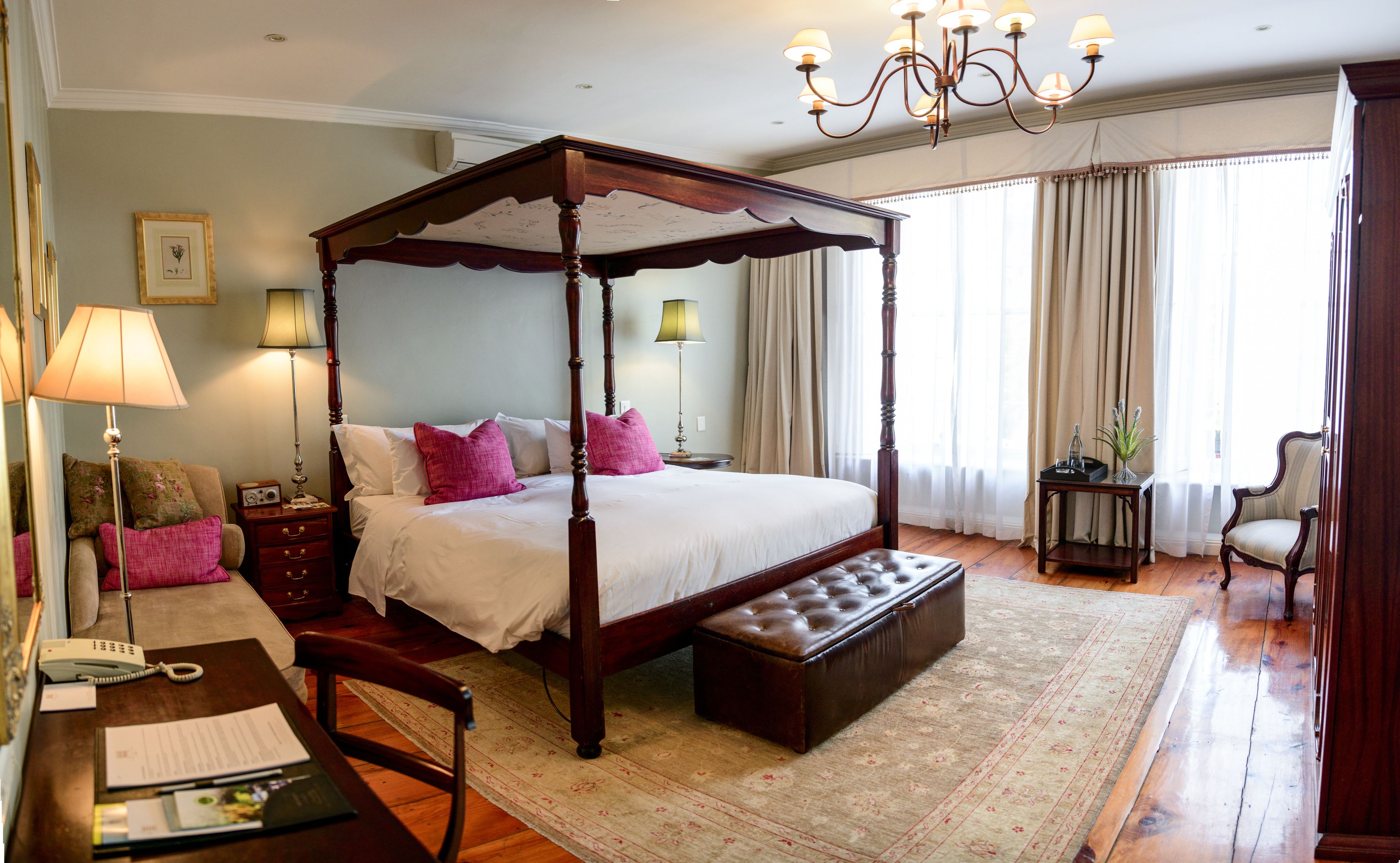 A four-poster bed with a bench, a desk with a telephone, and a chair in a room with wooden flooring and a large window with curtains.
