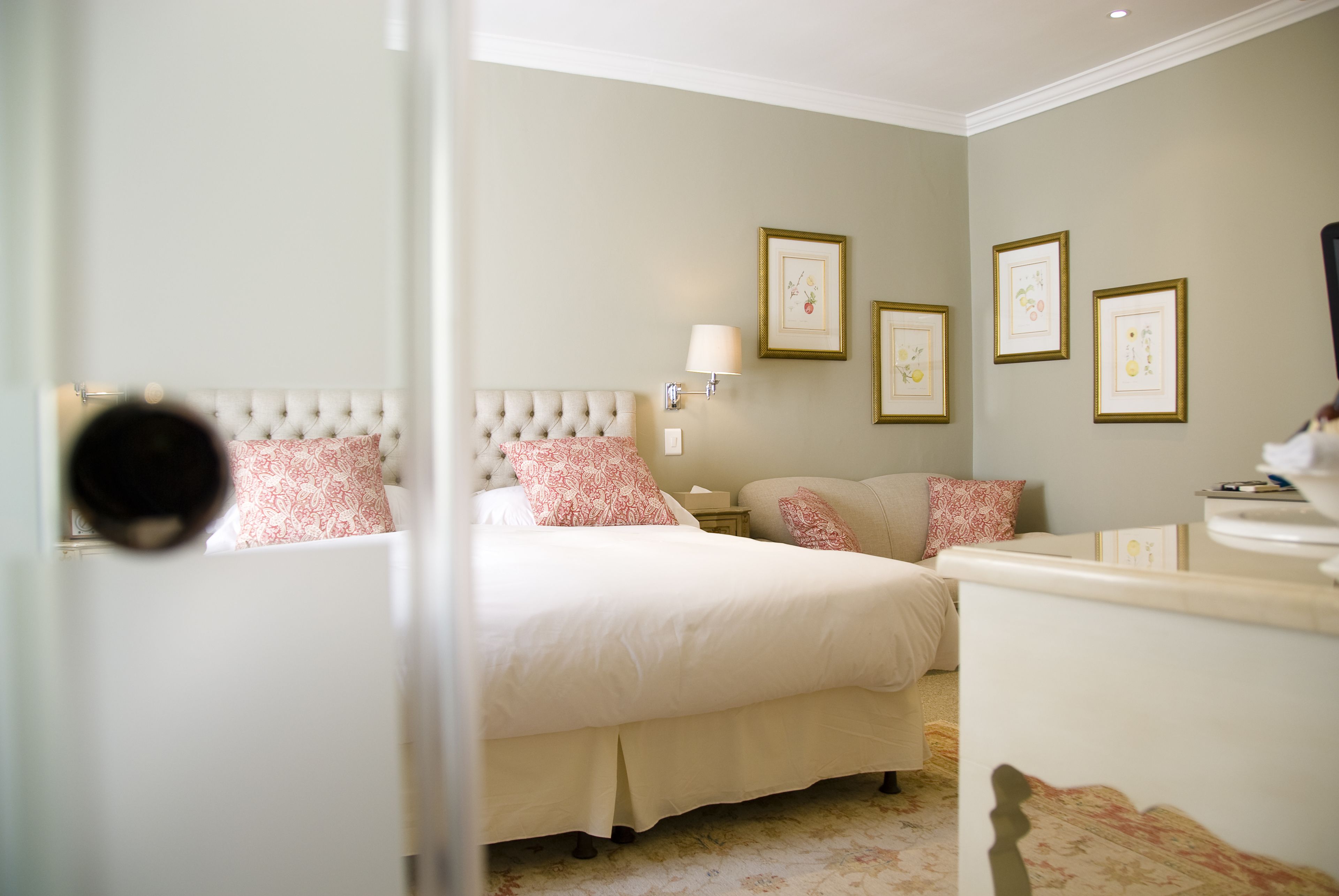 A bedroom with a tufted headboard, a bed with white and pink patterned bedding, a bedside table with a lamp, and framed pictures on the wall.