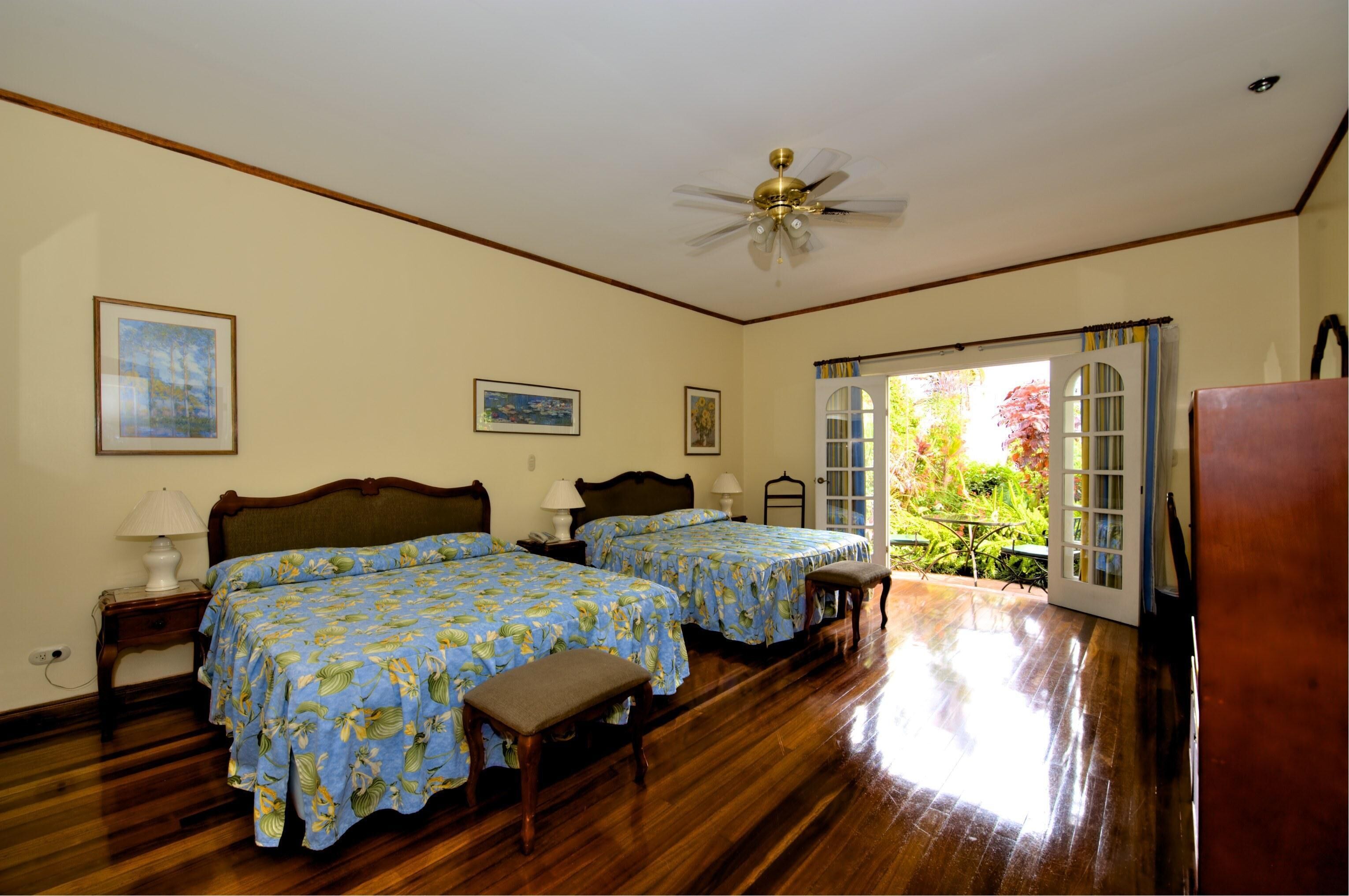 A bedroom with two beds, wooden flooring, a ceiling fan, and a view of the outdoors.