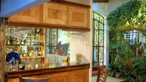 A bar area with wooden cabinets, shelves of liquor bottles, and hanging wine glasses. There are two wooden chairs and a round table. - Hotel Le Bergerac (San José)
