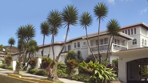 A hotel with a white facade, multiple balconies, and a well-maintained garden with palm trees. - Hotel Le Bergerac (San José)
