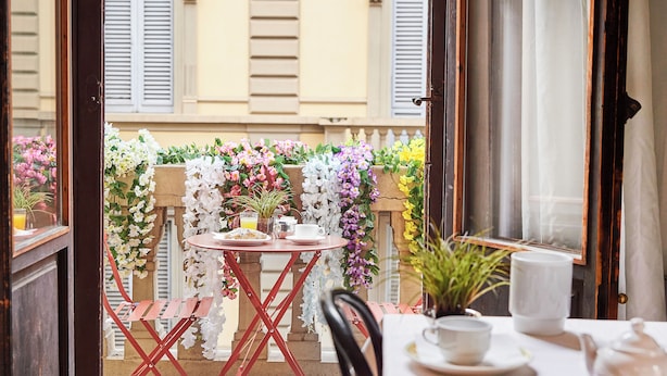 Un balcon avec une table préparée pour le petit-déjeuner, entouré de fleurs aux couleurs vives et offrant une vue sur un immeuble.
