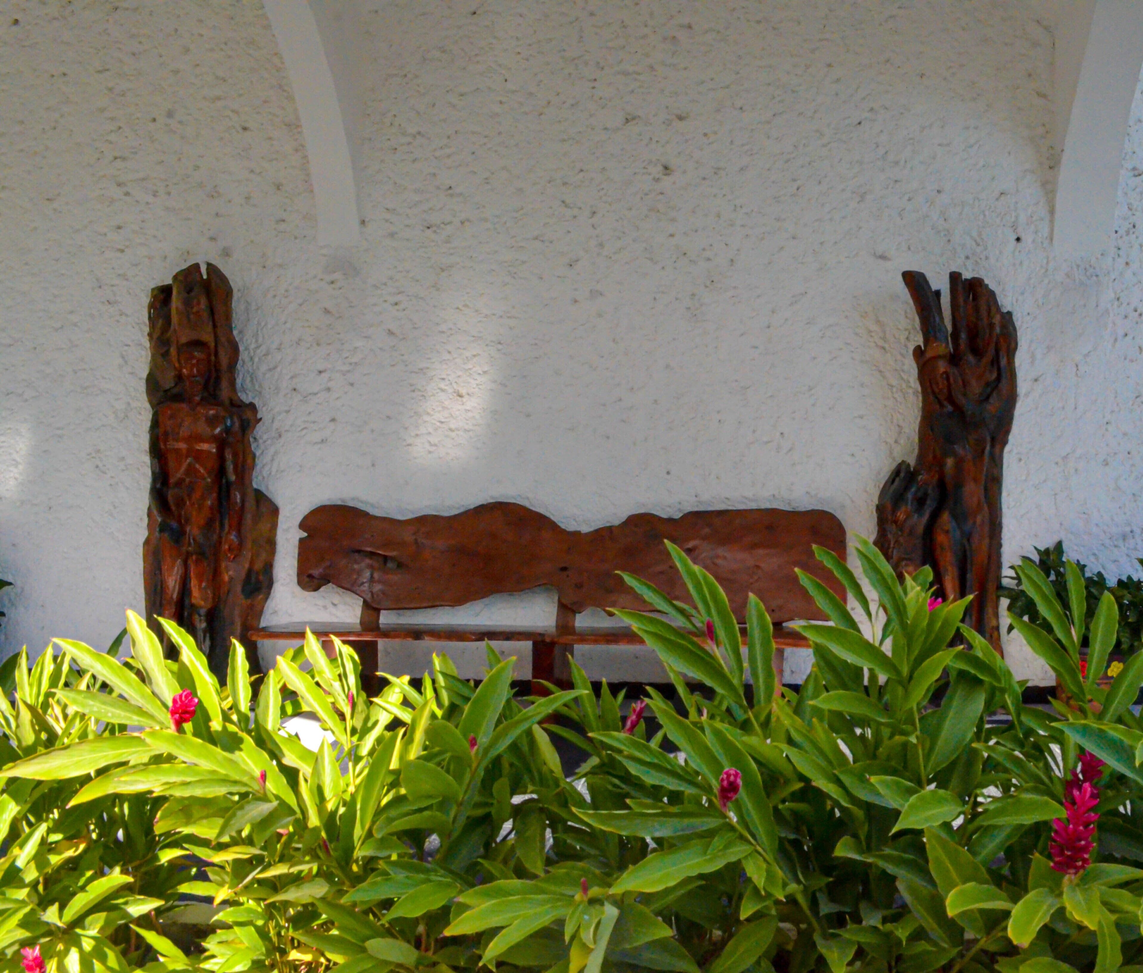 A wooden bench with carved figures on either side, set against a textured wall with green foliage in the foreground.