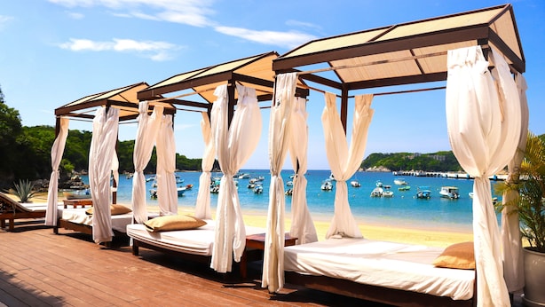 Una cabaña frente al mar con cuatro camas, cada una con dosel y almohadas, con vistas a una playa con barcos y un muelle.