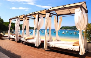 A beachside cabana with four beds, each with a canopy and pillows, overlooking a beach with boats and a pier.