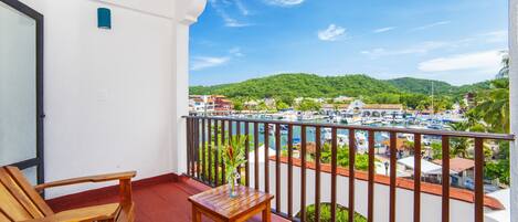 A balcony with wooden furniture, a small table, and a view of a marina with boats and a hilly landscape.