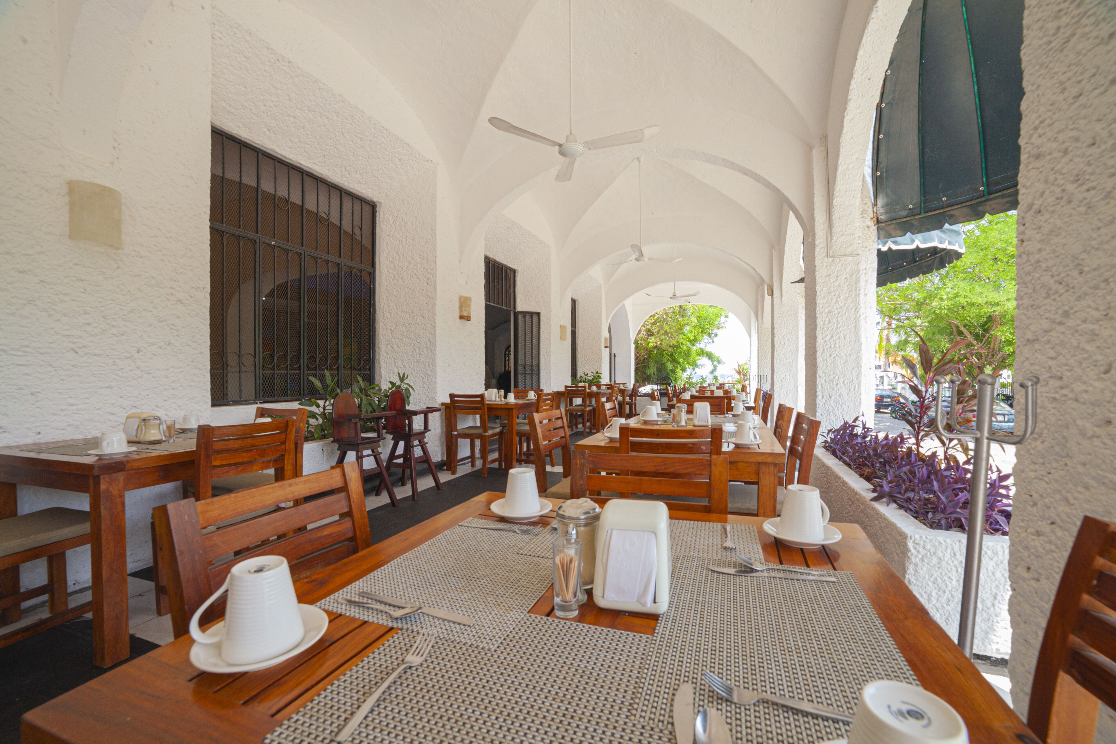 Uma ampla área de refeições interna com mesas e cadeiras de madeira, ventiladores de teto e grandes janelas que permitem a entrada de luz natural.