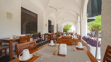 Uma ampla área de refeições interna com mesas e cadeiras de madeira, ventiladores de teto e grandes janelas que permitem a entrada de luz natural.