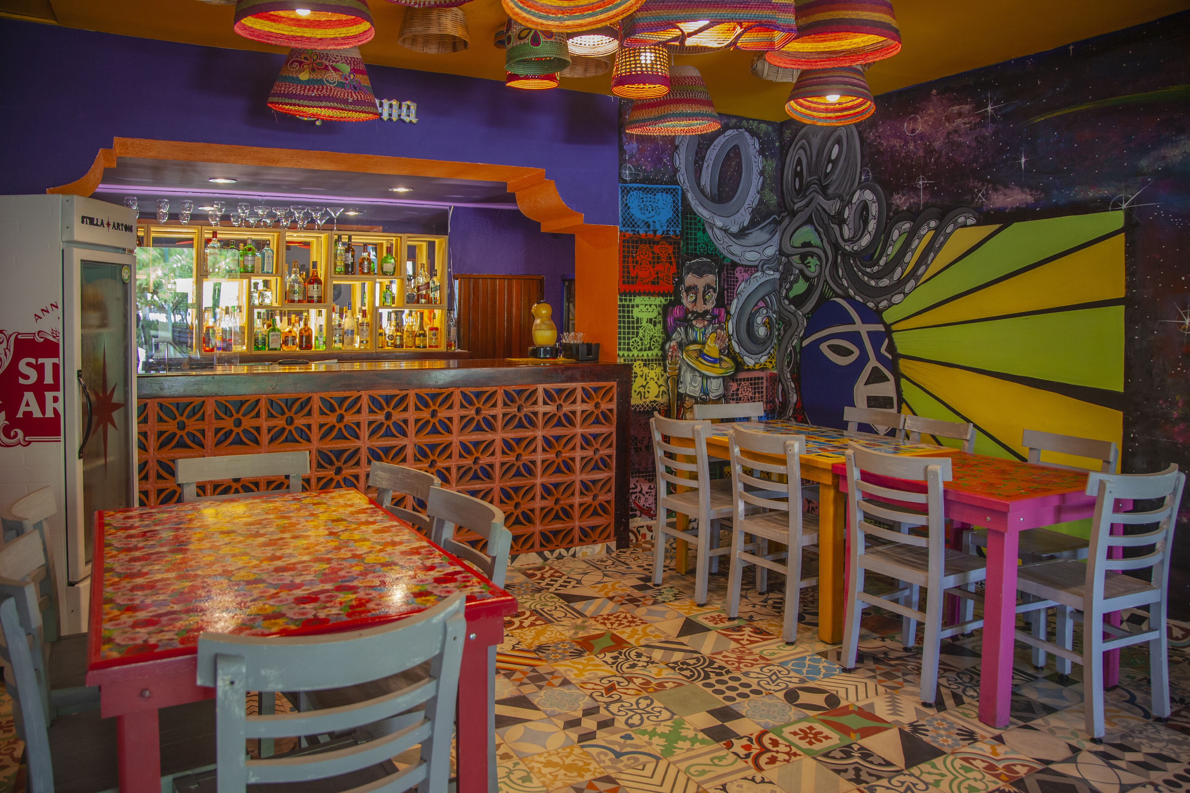 Interior vibrante de restaurante com área de bar, piso colorido com padrão e um mural na parede.