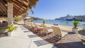 Terraza con sillones reclinables y plantas en macetas, con vista a una playa donde se ven un crucero y varias embarcaciones.