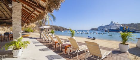 A deck with lounge chairs and potted plants, overlooking a beach with a cruise ship and boats.