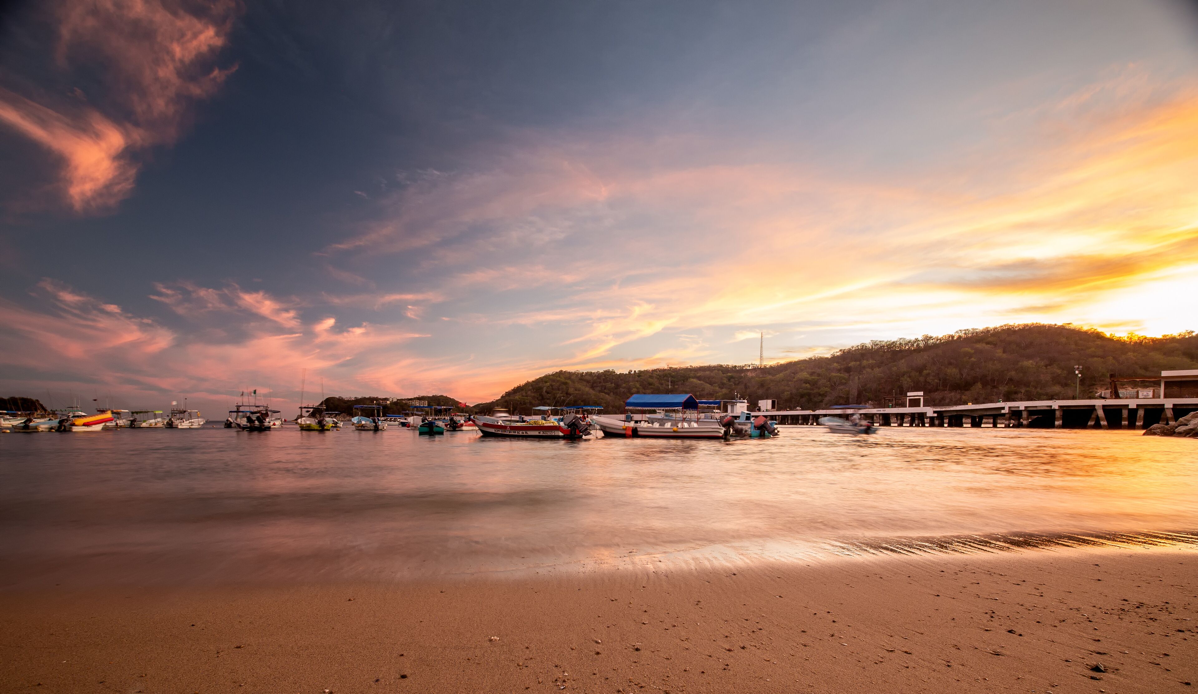 A waterfront scene with boats docked at a pier, a sandy beach in the foreground, and a hilly landscape in the background.