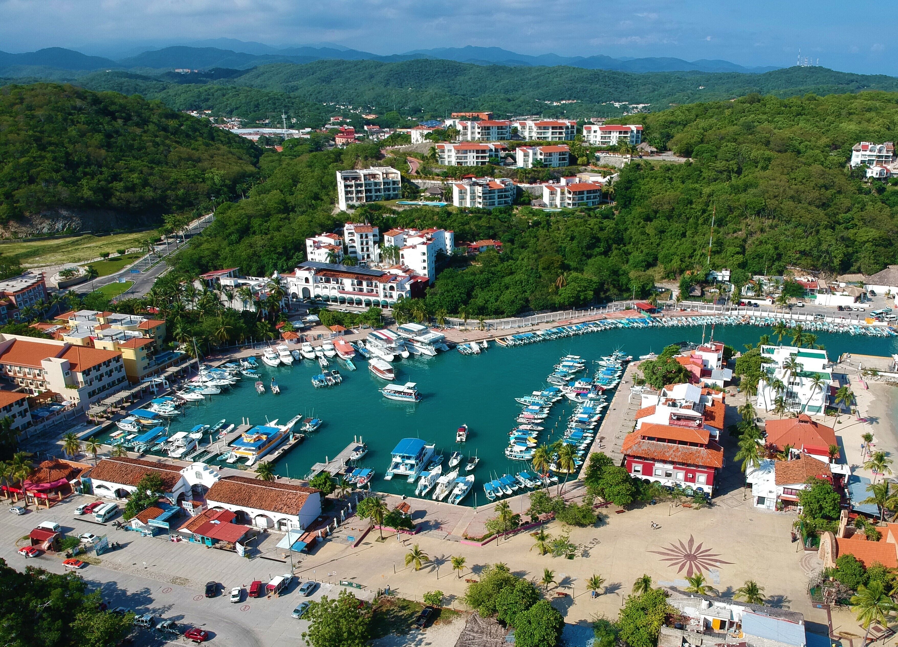 A marina with numerous boats docked, surrounded by buildings with red roofs and a mountainous backdrop.