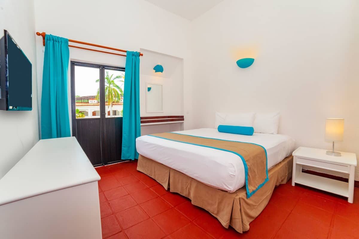 Een hotelkamer met een bed, nachtkastje, televisie, raam met gordijnen en uitzicht op palmbomen.