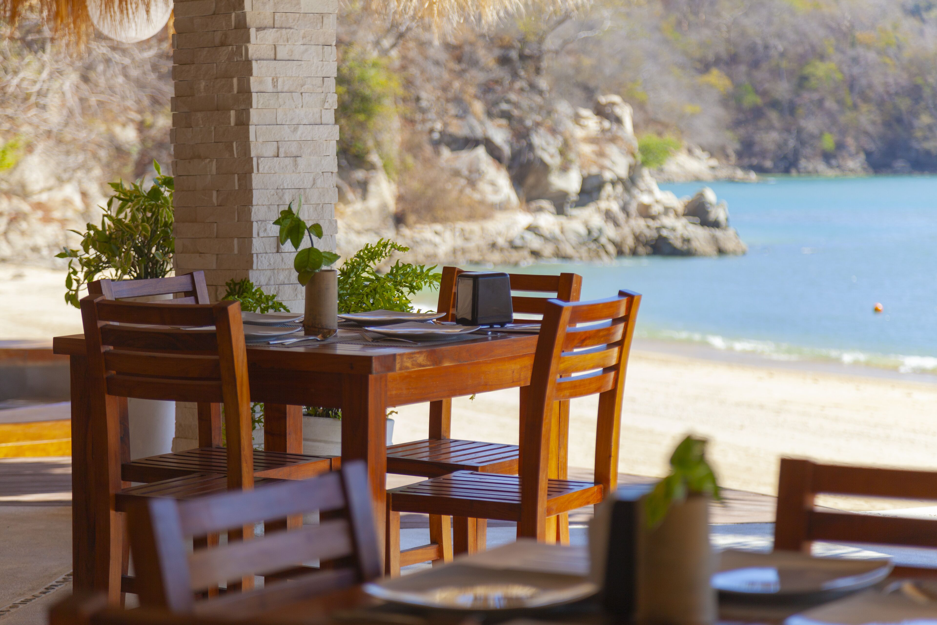 Une table en bois, entourée de chaises et sur laquelle est posé un ordinateur portable, offre une vue sur une plage bordée de falaises rocheuses et sur une mer calme.