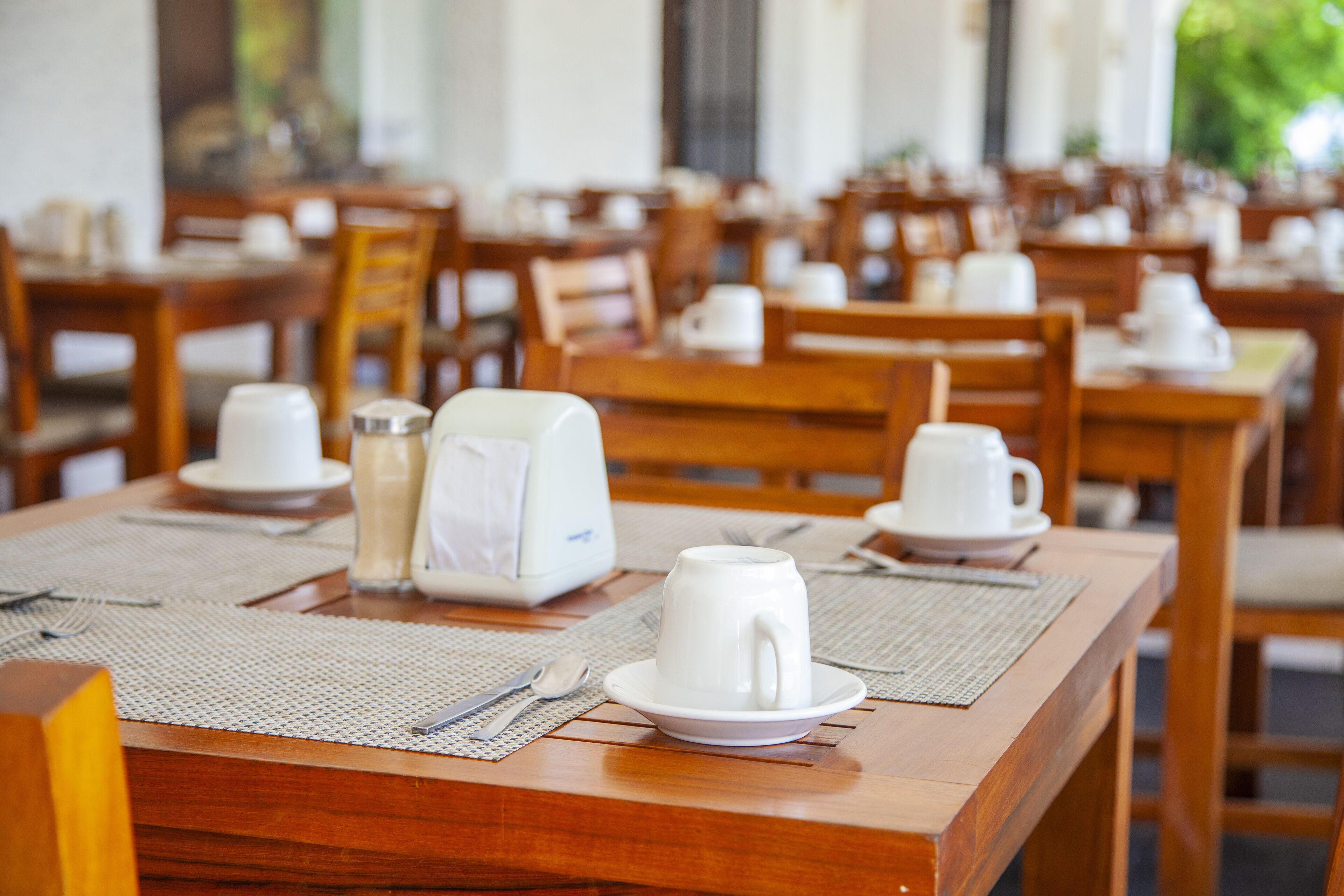 Comedor con mesas y sillas de madera, vajilla blanca y una cafetera.