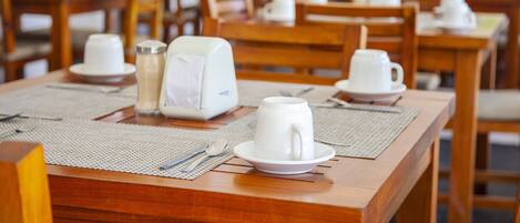 Comedor con mesas y sillas de madera, vajilla blanca y una cafetera.