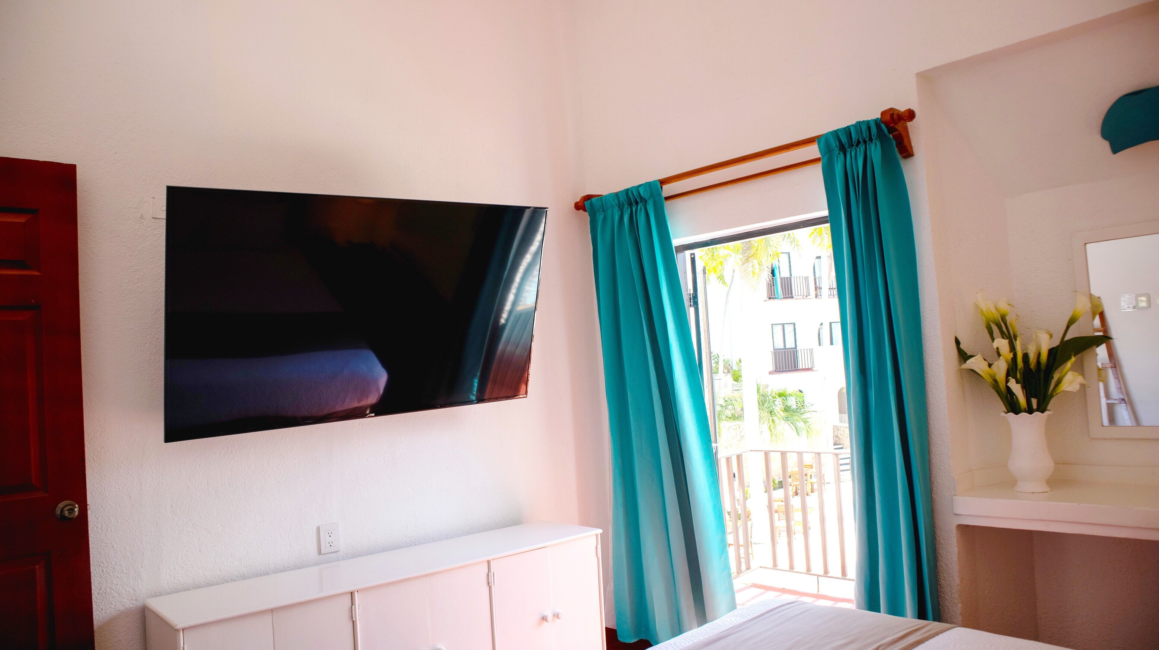 A hotel room with a flat-screen TV mounted on the wall, a white dresser, a bed, and teal curtains.