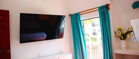 A hotel room with a flat-screen TV mounted on the wall, a white dresser, a bed, and teal curtains.