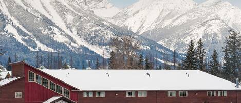 A large building with a red facade and a snow-covered roof, surrounded by a parking lot and mountains in the background.