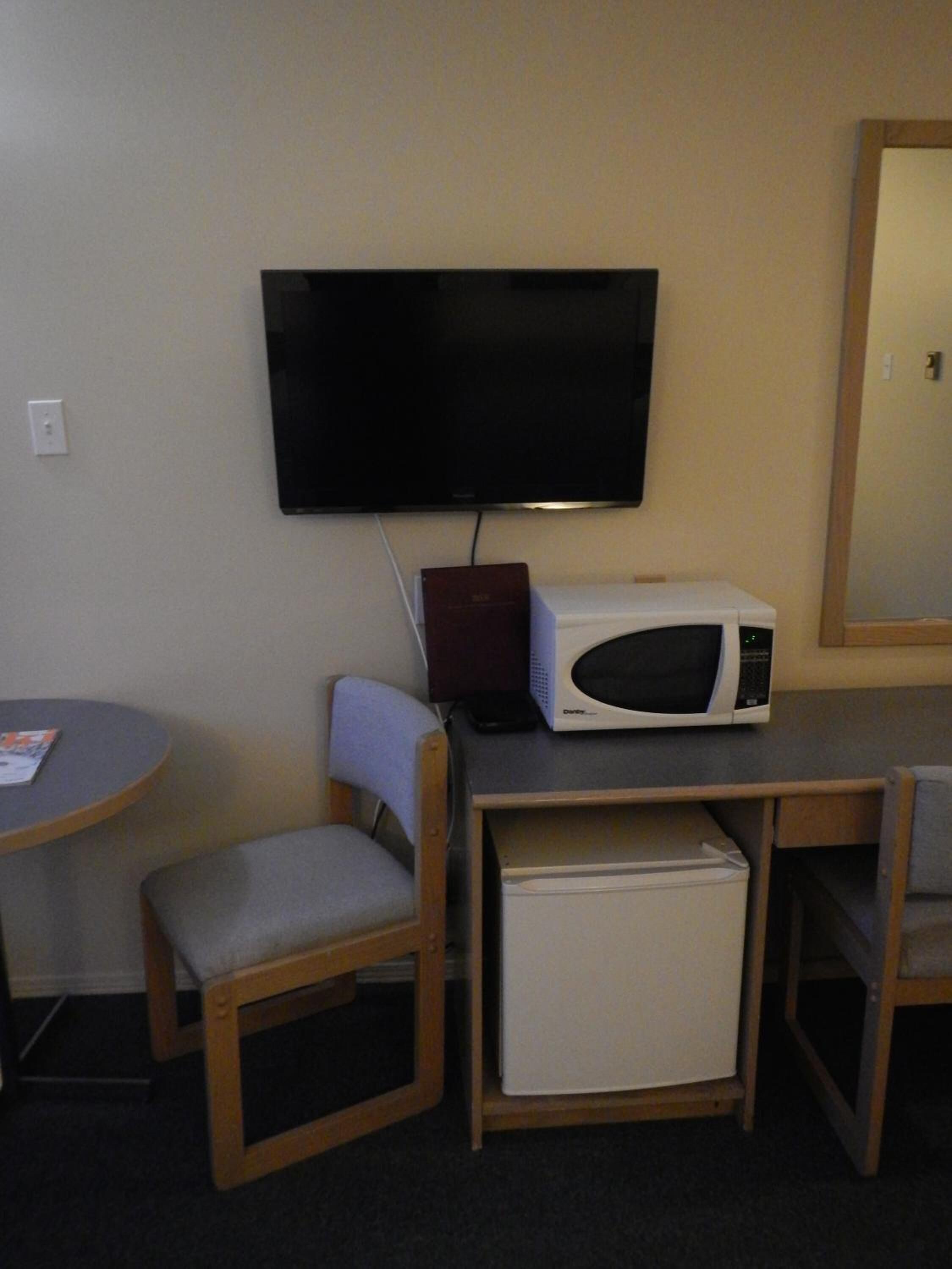 A compact room with a flat-screen TV mounted on the wall, a microwave, a small refrigerator, and a chair.