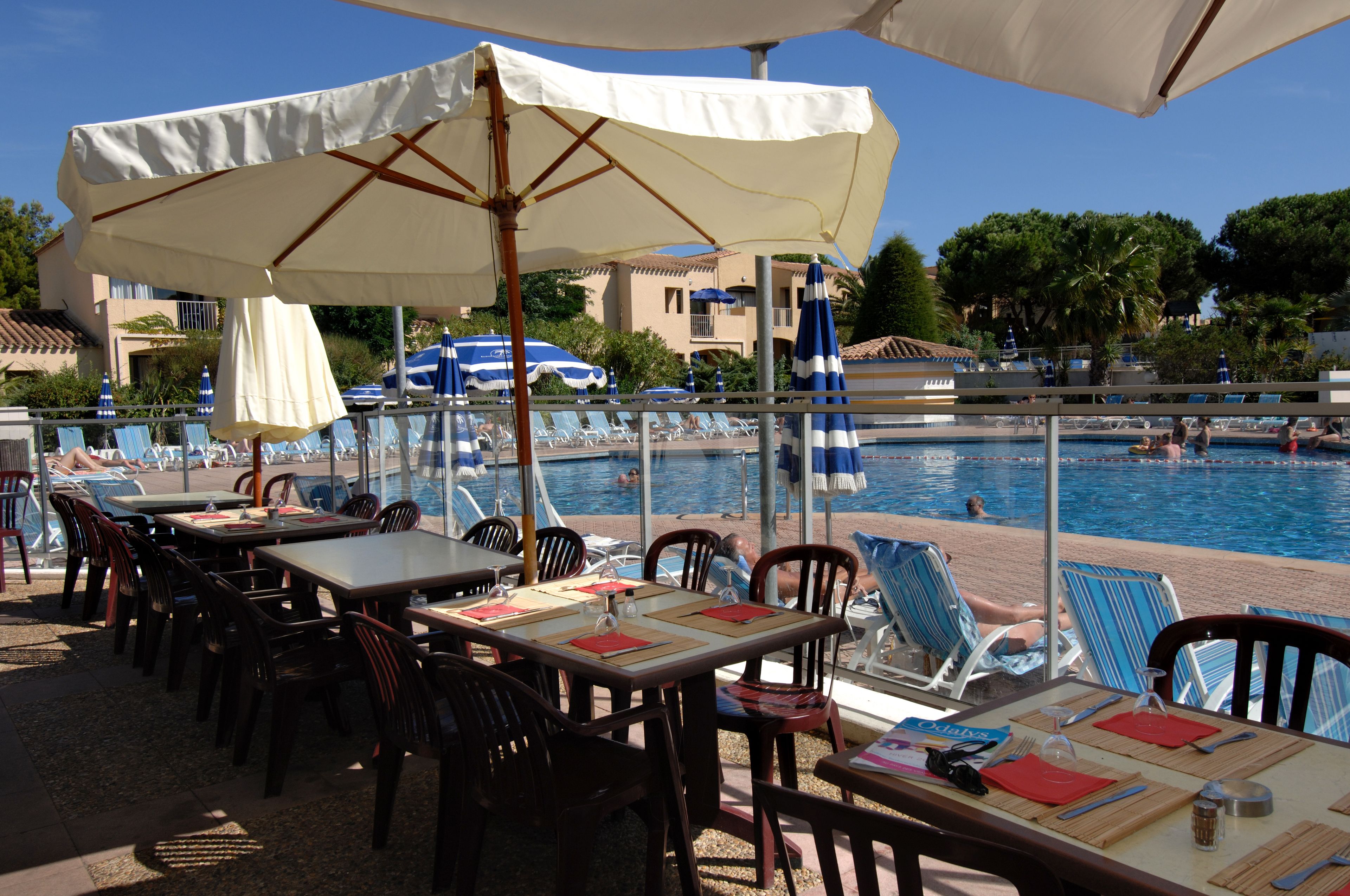 Un espace aménagé au bord de la piscine, avec des tables et des chaises sous des parasols, donnant sur une piscine.