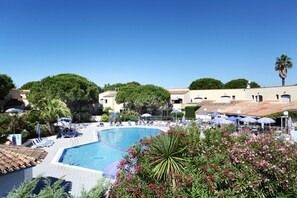 A pool area with lounge chairs and umbrellas, surrounded by greenery and buildings with tiled roofs. - Odalys Résidence Saint Loup (Agde)