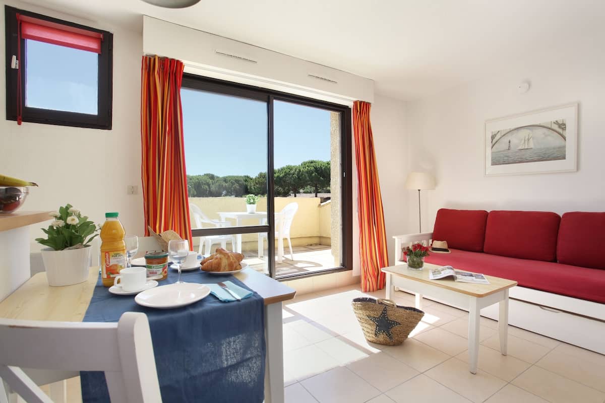 Un salón luminoso con un sofá rojo, una mesa de comedor con alimentos para el desayuno y vistas al exterior.