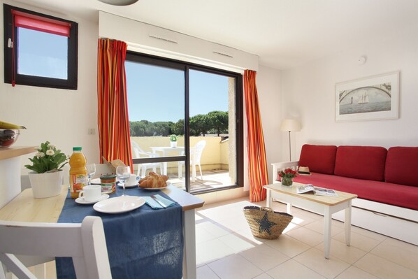 A bright living room with a red sofa, a dining table with breakfast items, and a view of the outdoors. - Odalys Résidence Saint Loup (Agde)