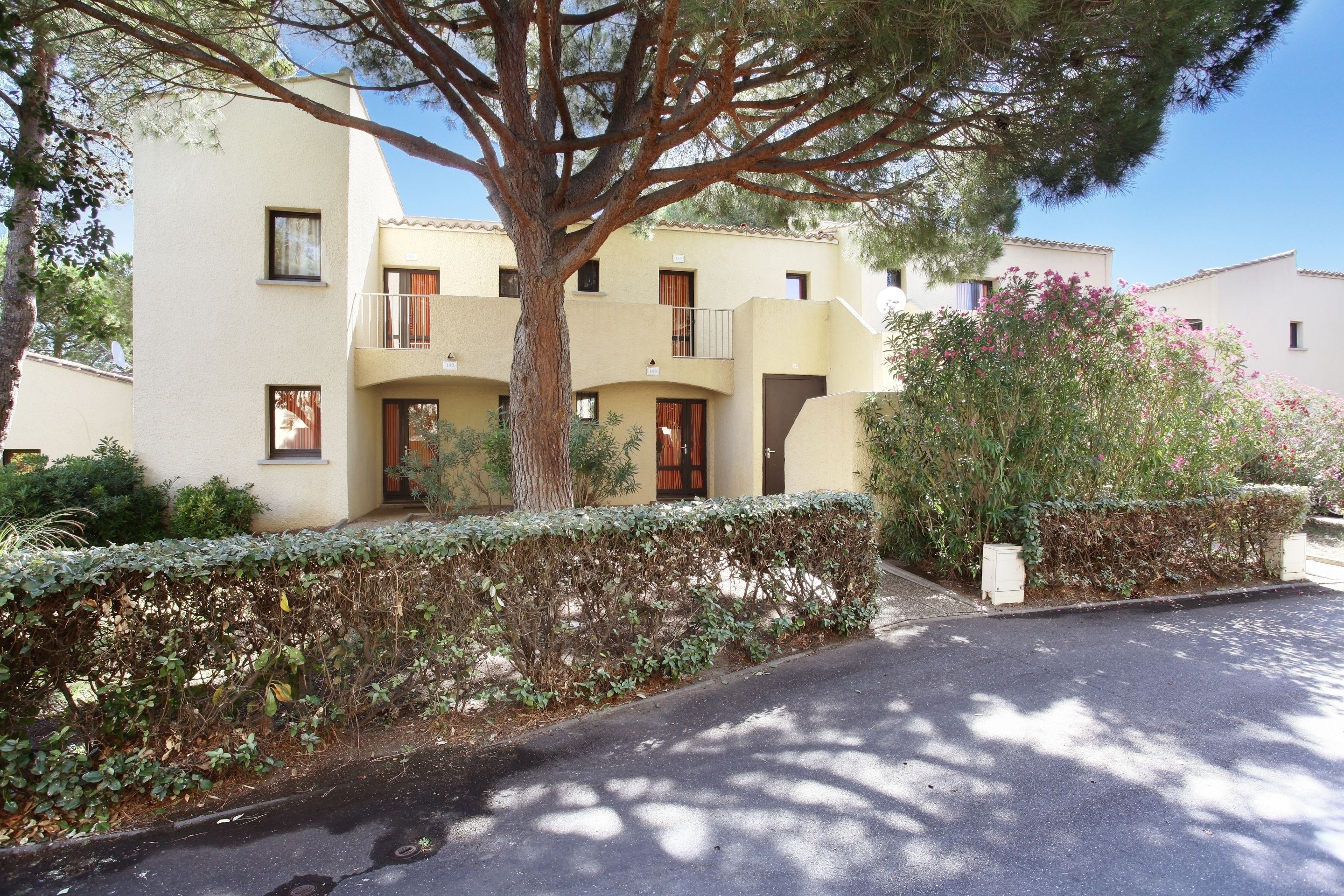 Un edificio a più piani con balconi, un albero di fronte e un siepe lungo il marciapiede.