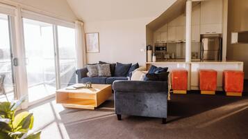 Una sala moderna con sofá, reposapiés y mesa de centro, un balcón con puerta corrediza, y una cocina con armarios y electrodomésticos.