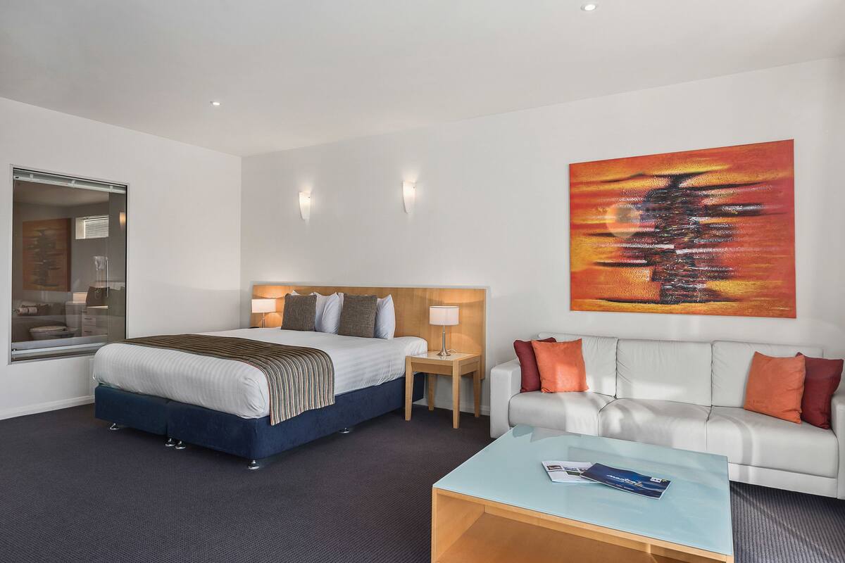 A hotel room with a bed, a bedside table with a lamp, a sofa, a coffee table, and a painting on the wall.