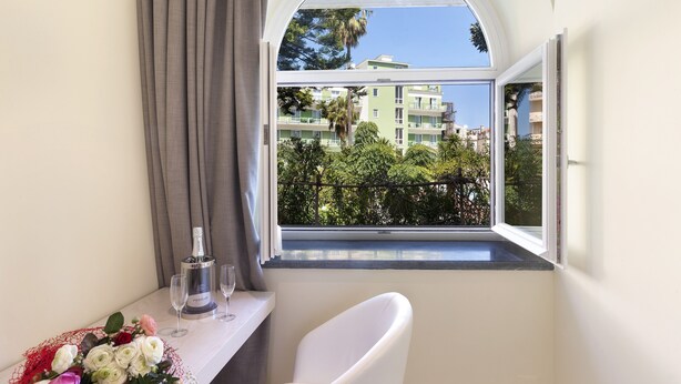Ein Zimmer mit einem großen Fenster, einem weißen Stuhl, einem Tisch mit Flasche und Gläsern sowie einem Blumenstrauß.